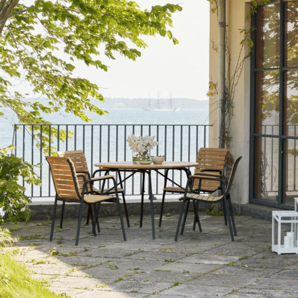 A MANDALAY – Magnolia & Bellevue Havesæt Ø110 (Kopier) med rundt træbord og fire tilhørende stole er indrettet på et stenbelagt areal. Terrassen har udsigt over et vandområde med høje skibe i det fjerne. En glasdør og potteplanter er synlige i baggrunden.