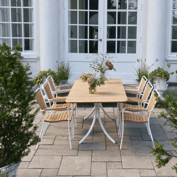 A MANDALAY – Magnolia & Marguerit Havesæt L197 med en vase med blomster på toppen er omgivet af seks matchende træ- og metalstole. Opsætningen er på en stenbelagt terrasse foran en hvid bygning med store glasdøre og vinduer. Grønne planter er placeret rundt om terrassen.