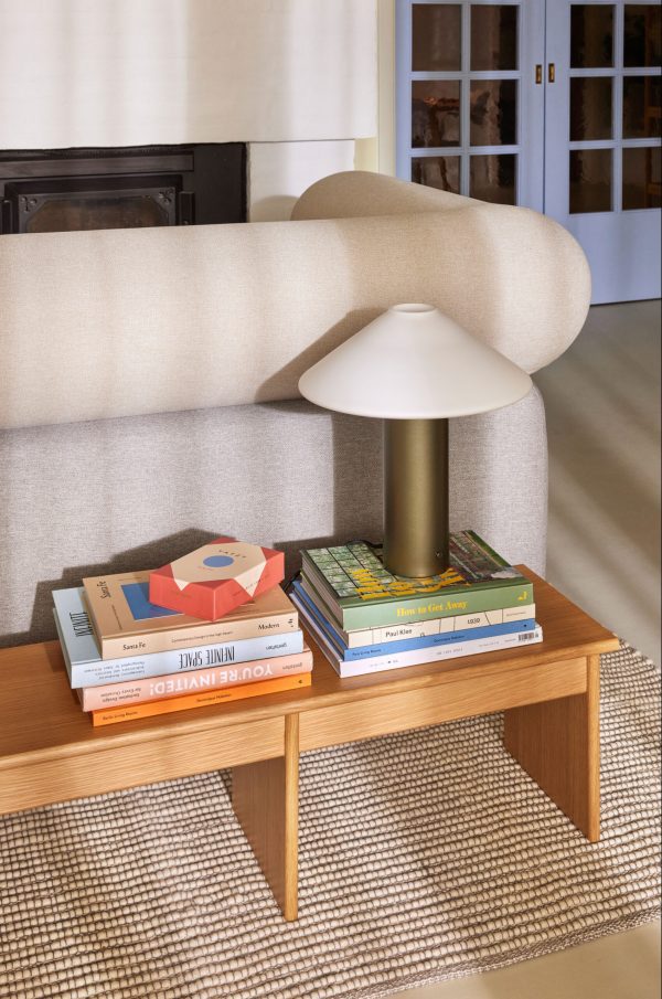 Et hyggeligt stuehjørne med en moderne lampe på et sofabord i træ stablet med farverige bøger, sat op mod et tekstureret tæppe og en beige sofa.