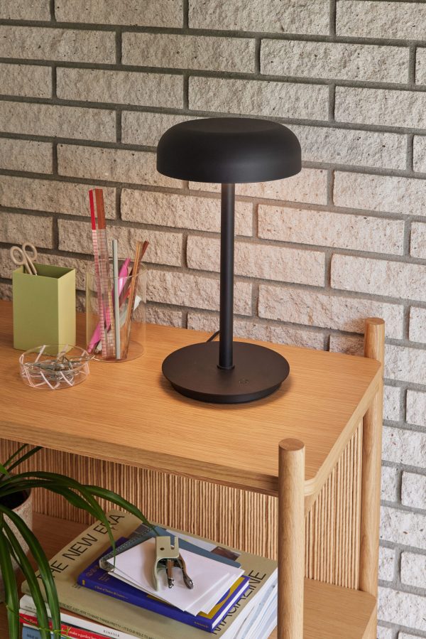 En moderne sort skrivebordslampe står på en bogreol i træ, som rummer blyanter, notesblokke og bøger. hylden er sat på baggrund af hvid murstensvæg.
