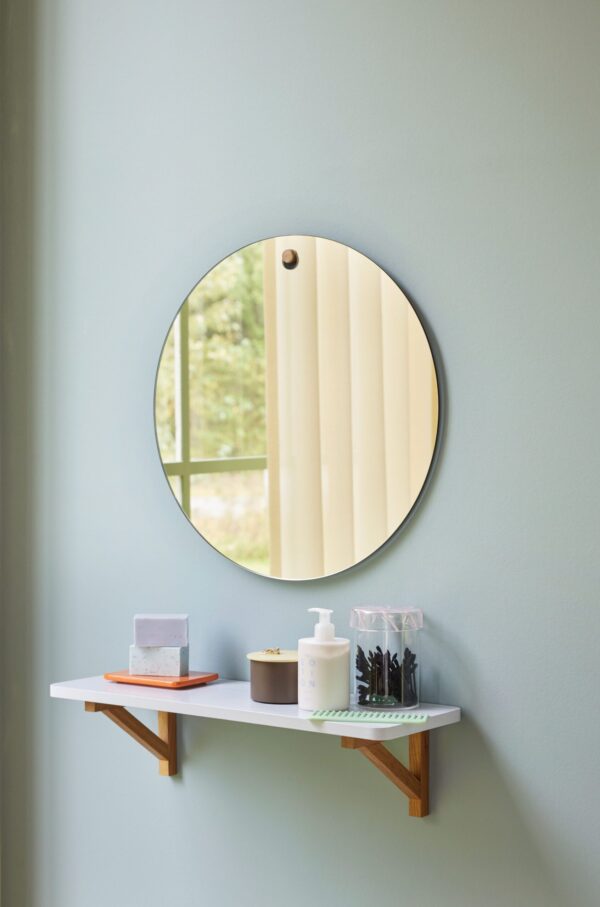 Et cirkulært spejl hænger på en lyseblå væg over en hvid hylde med toiletartikler og makeupartikler. spejlet reflekterer et vindue, der viser dagslys og træer.