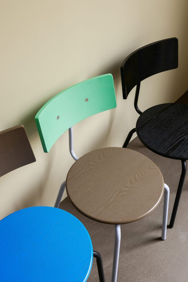 En samling af unikke stole og borde med forskellige farver og designs; inkluderer et bord med blåt bord og en stol med grøn ryg, mod en beige væg.