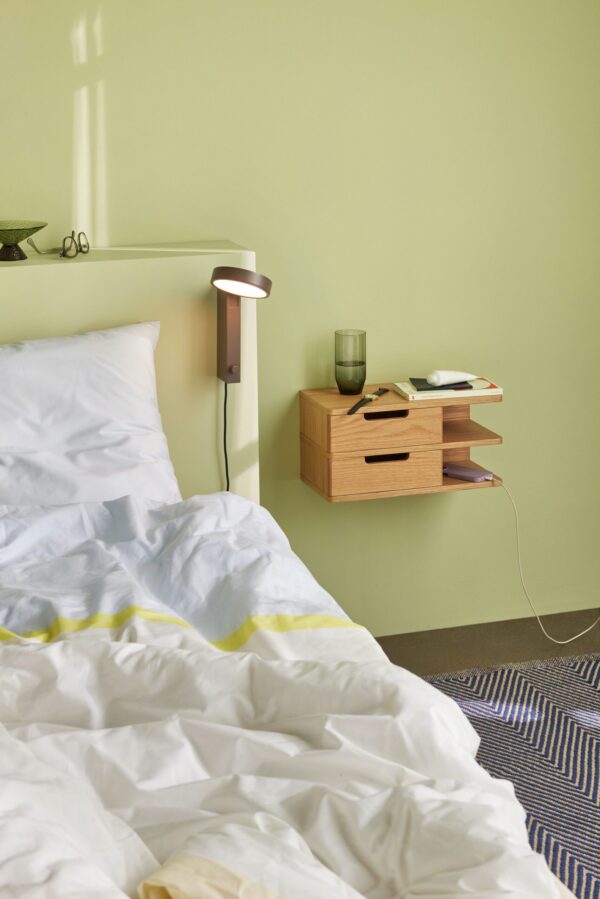 En pæn opredt seng med hvidt og gult sengetøj i et rum med grønne vægge. Et natbord af træ, der viser TomorrowsDesign, rummer en lampe, bøger, et glas vand og en smartphone. Lyset strømmer blødt ind fra venstre.