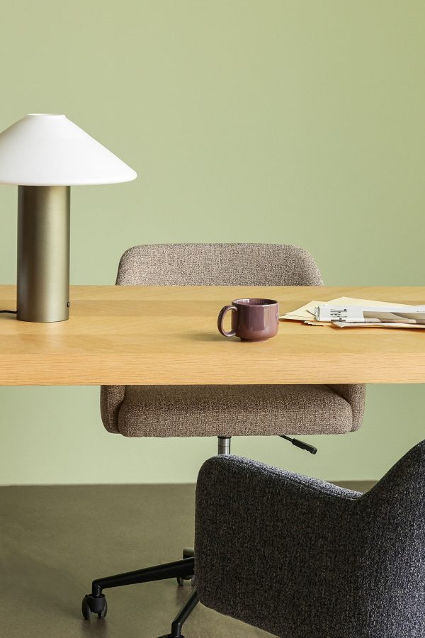 Et velorganiseret arbejdsområde med et træskrivebord med et brunt krus, spredte papirer og en lampe. en polstret stol er gemt under skrivebordet, sat op ad en tofarvet grøn væg.