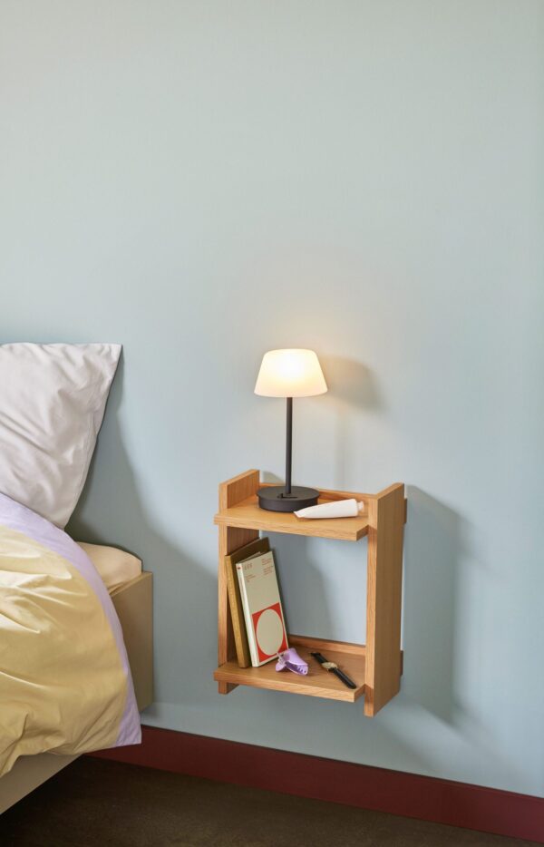 Et lille sengebord af træ med en oplyst lampe, en bog og et par briller placeret på den nederste hylde, ved siden af en seng med hvidt og gult sengetøj mod en lyseblå væg.