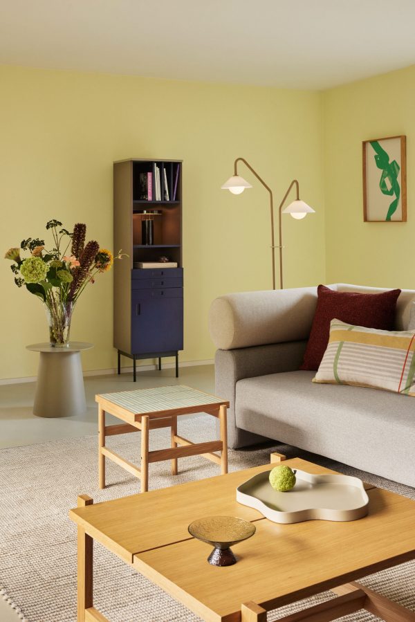 En hyggelig stue med en beige sofa, et sofabord i træ og et sidebord med en vase med blomster. et lille skab og et grønt maleri pryder de gule vægge, suppleret med en gulvlampe.