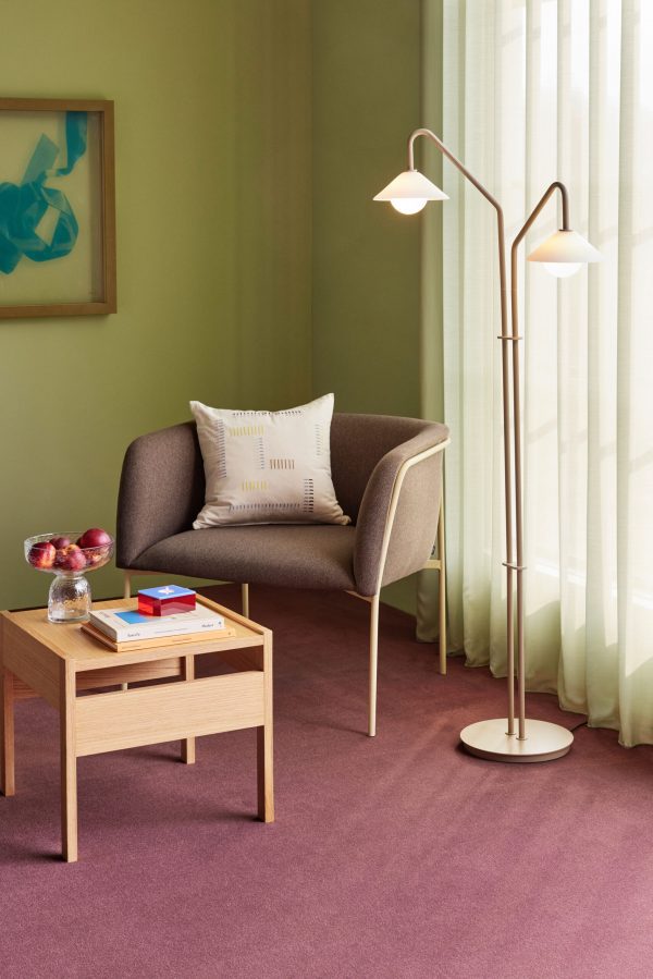 En hyggelig læsekrog med en grå lænestol, et sidebord i træ med bøger og en skål frugt og en høj, moderne gulvlampe med to lys. rummet har grønne vægge og et stort vindue med gennemsigtige gardiner.