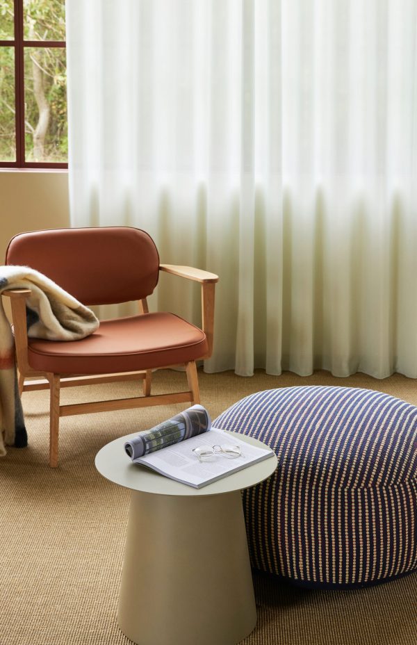 Interiør af et værelse med en terracotta-farvet stol med et beige tæppe, et hvidt sidebord med et magasin og en stribet blå puf på et beige tæppe, nær rene hvide gardiner med udsigt til haven.