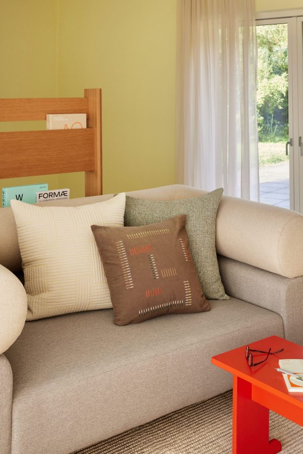 Et hyggeligt stuehjørne med en beige sofa med diverse pudepuder i jordfarver. et lille rødt sidebord, grønne gardiner og udsigt til haven gennem en glasdør fuldender scenen.