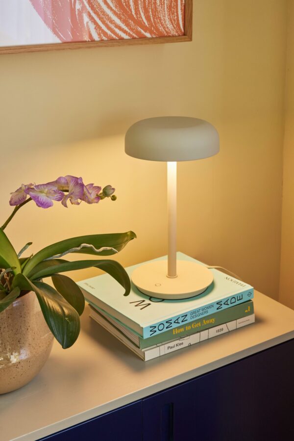 En moderne bordlampe med en grå skærm og hvid base står på en stak bøger ved siden af en blomstrende lilla orkidé, mod en væg malet i en gradient fra gul til orange.