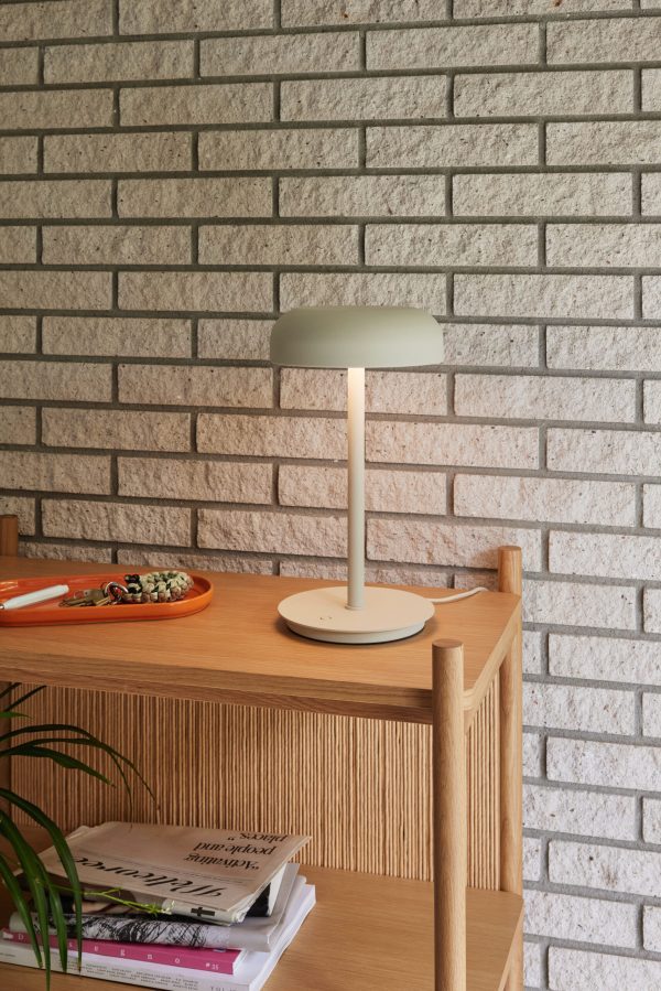 En moderne lampe med en dæmpet grøn skærm og hvid base står på et træbord mod en murstensvæg. bordet rummer også magasiner og en tallerken med snacks.