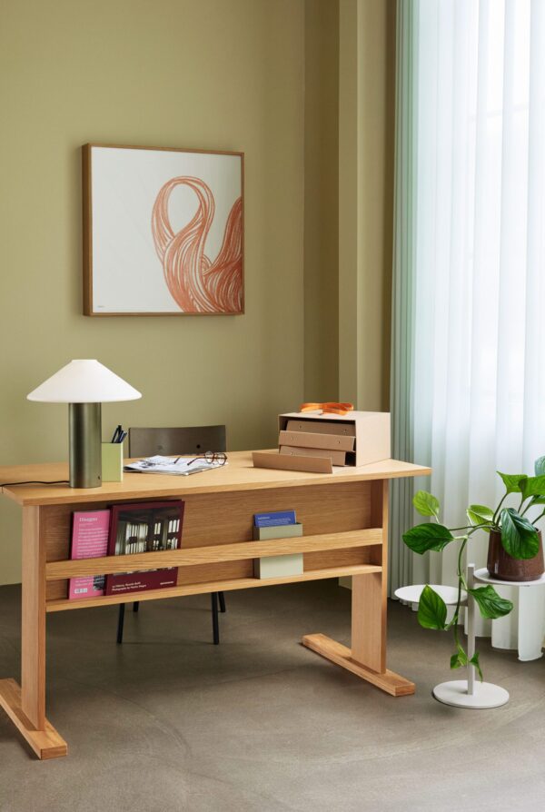 Et minimalistisk hjemmekontor med et træskrivebord med bøger, en lampe og papirvarer. der er et indrammet abstrakt kunstværk på væggen og en potteplante ved siden af skrivebordet. lyset filtrerer gennem rene gardiner.