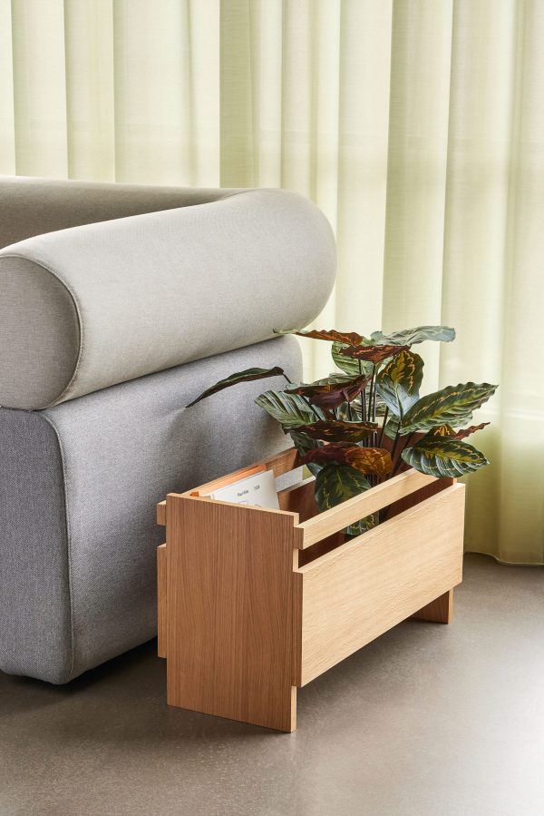 Et moderne stuehjørne med en lysegrå sofa med et afrundet armlæn og et sideplantestativ i træ med grønne potteplanter. lysegrønne gardiner giver en blød baggrund.