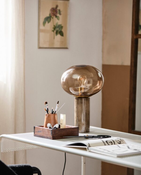 Et moderne, minimalistisk arbejdsområde med et elegant hvidt skrivebord med en unik overdimensioneret pærebordlampe på en rustik base. en trækasse rummer forskellige kunstbørster, ved siden af åbne notesbøger og læsebriller.