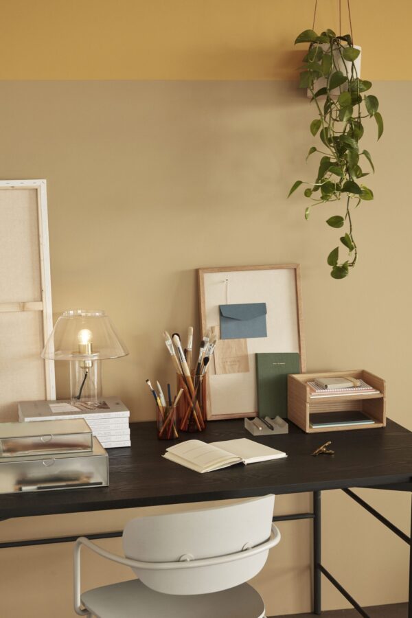 Et ryddeligt arbejdsområde med et sort skrivebord med en Hübsch Arch spisestuestol lysegrå. Skrivebordet rummer diverse papirvarer, en lampe med en klar glasskærm, notesbøger, planlæggere og pensler. En potteplante hænger på den beige og gule væg i baggrunden.