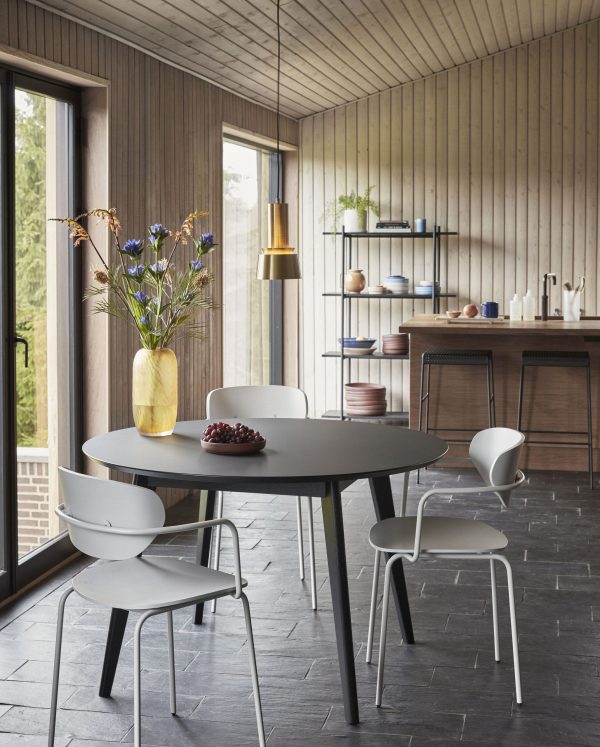 En moderne spiseplads med et rundt sort bord og fire Hübsch Arch Spisestuestole i lysegrå. En vase med blomster står på bordet sammen med en skål frugt. Rummet har trævægge, store vinduespartier og udsigt til grønt udenfor. Reoler med tallerkener er synlige i baggrunden.