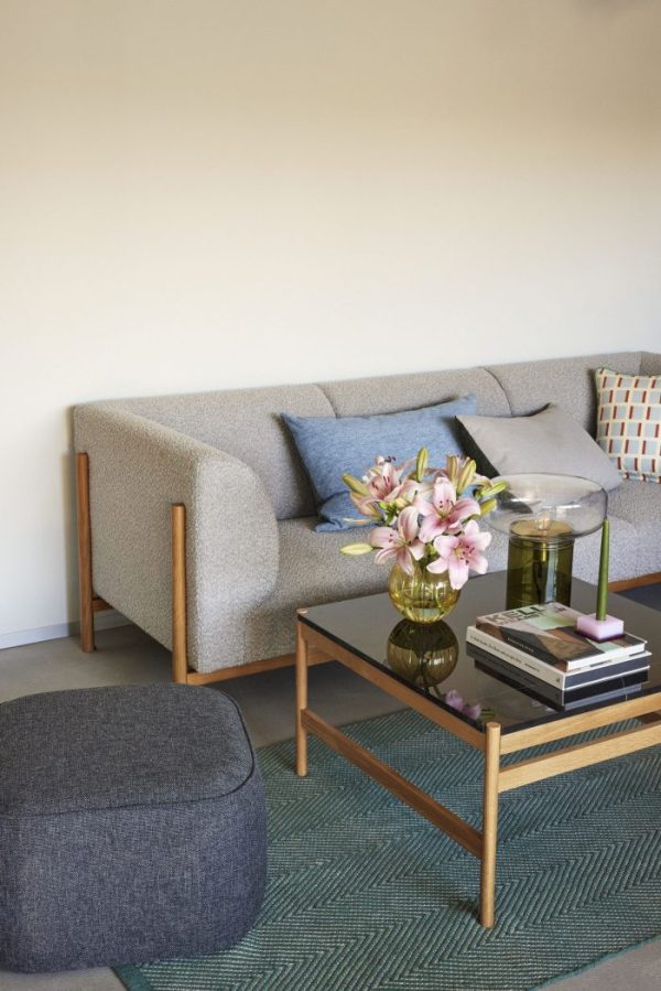 En moderne stue har en HÜBSCH - Moment Sofa 3-pers. (Kopier), en blå og en mønstret pude, et sofabord med glasplade med bøger, et lys og en vase med lyserøde blomster. En grå skammel og et blågrønt tæppe er også synlige. Baggrunden er en neutral-tonet væg.
