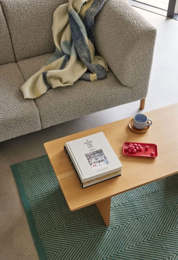En moderne stuescene med en HÜBSCH - Moment Sofa 3-pers., et tæppe draperet over armen, et sofabord i træ med stablede bøger, en tallerken med røde bær og en kop og underkop. Bordet står på et grønt mønstret tæppe, og der er udsigt ud til en altan.