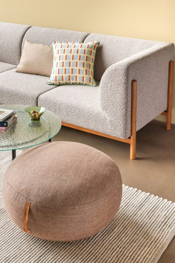 En moderne stue har en HÜBSCH - Moment Sofa 3-pers. med træben, en geometrisk mønstret pude, en rund beige puf, et sofabord i glas med en stak bøger og et lille fad og et neutralt tonet tæppe på et gråt gulv.