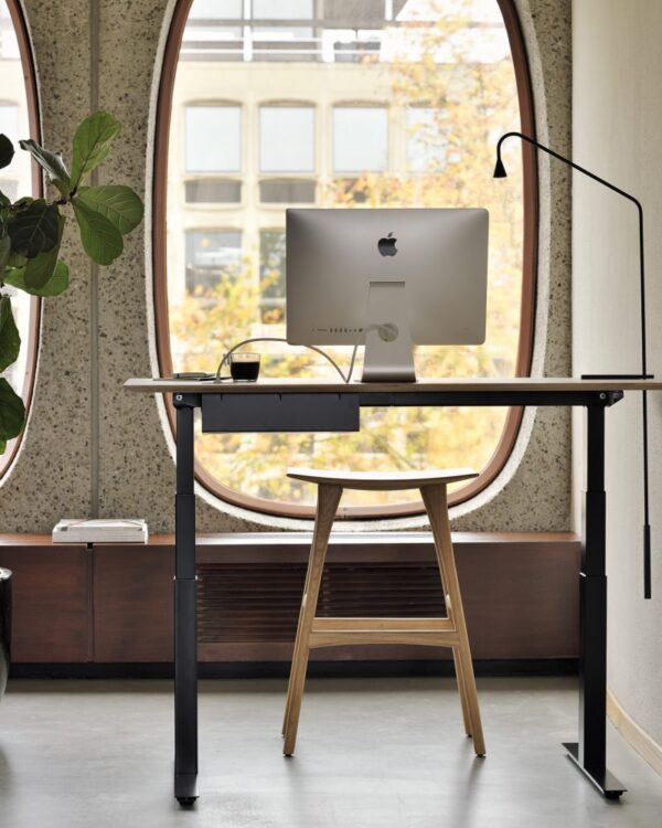 Et moderne arbejdsområde har et elegant Bok justerbart skrivebord - lakeret egetræsplade - sort base - rektangulær - med kabelstyring - EU med en træstol, en Apple stationær computer og en sort skrivebordslampe. Skrivebordet er placeret tæt på store, bueformede vinduer med udsigt til en bygning og efterårstræer udenfor. En potteplante er delvist synlig til venstre.
