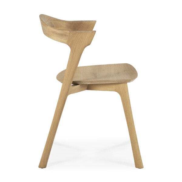 En Bok spisestuestol - eg med minimalistisk design er vist i en sideprofil. Stolen har buet ryglæn og sæde, begge lavet af lyst træ, understøttet af tre vinklede ben. Designet er moderne og slankt og fremhæver den naturlige træåre.