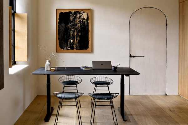 Et minimalistisk hjemmekontor har et Bok-justerbart skrivebord - sort lakeret eg - sort base - rektangulær - med kabelstyring - EU med en bærbar computer, gren i en hvid vase, fad og en kop. Tre trådstole står ved skrivebordet. Et abstrakt maleri i en træramme hænger på en lys væg med en buet dør. Lyset strømmer gennem et vindue.