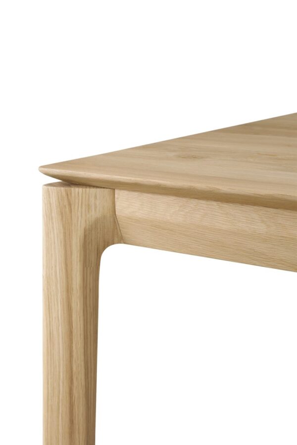 Dette billede viser et nærbillede af hjørnet af et Bok-skrivebord - lakeret eg - rektangulært - med kabelstyring. Materialet fremstår som lyst træ med en glat finish. Bordets design har rene linjer og en minimalistisk stil.