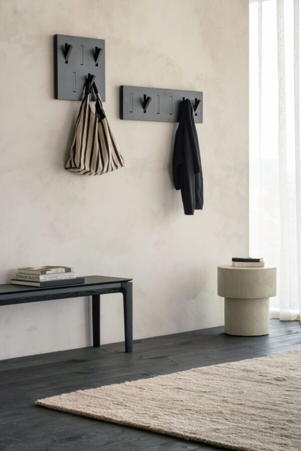 Et minimalistisk, lyst værelse med to sorte væghængte kroge, der holder en stribet taske og en mørk frakke. En enkel Bok-bænk - lakeret eg - sort med bøger på står op ad væggen. En beige cylindrisk skammel og stak bøger er tæt på et gennemsigtigt gardin. Et neutralt tæppe ligger på det mørke gulv.