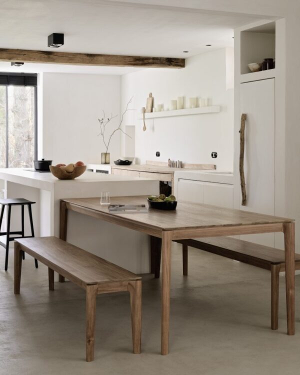 Et minimalistisk køkken og spiseplads med træmøbler. Spisebordet og matchende Bok bænk - teaktræ er lyst træ. Hvide vægge og kabinetter suppleres af moderne indretning, herunder planter og stearinlys. Et stort vindue giver naturligt lys.