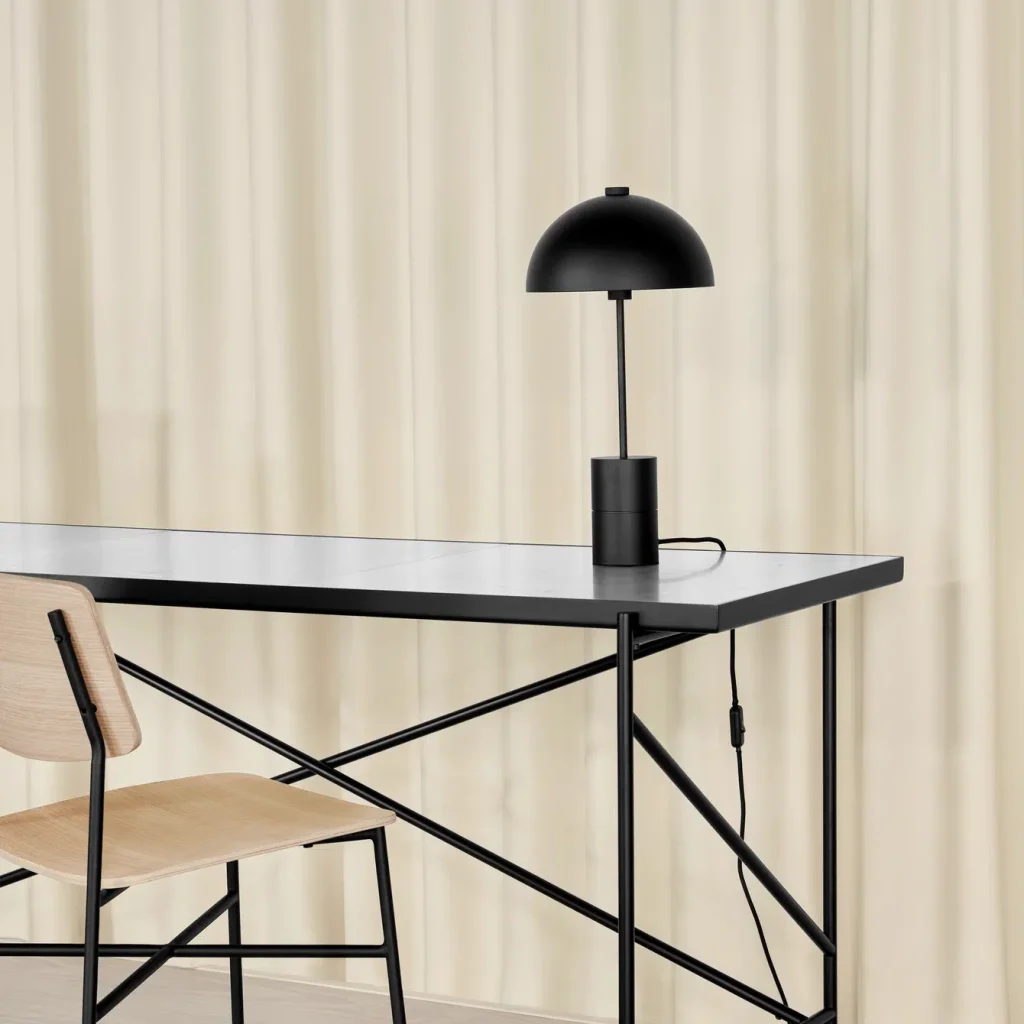 En minimalistisk hjemmekontoropsætning med et elegant sort skrivebord med metalben, en træstol på siden og en sort kuppellampe på skrivebordet, sat mod en blød beige gardinbaggrund.