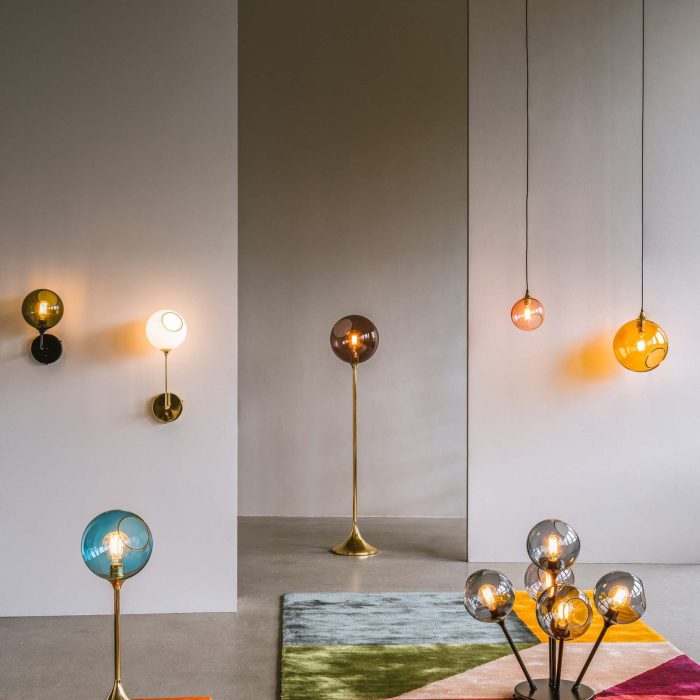 Et indvendigt rum med forskellige DESIGN BY US - Ballroom Lampe XL, inklusive gulv- og vægmonterede modeller. lamperne har forskellige designs, og rammerne omfatter farverige tæpper, der skaber en varm, kunstnerisk atmosfære.