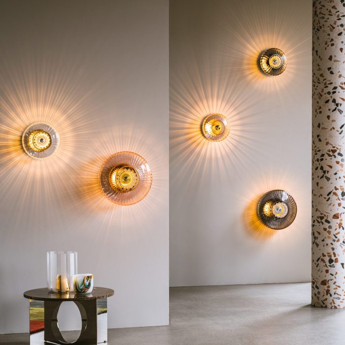 Et moderne stuehjørne med DESIGN BY US - New Wave Optic Væglampe XL, der støber varme lysmønstre på en grå væg. et lille, flerfarvet sidebord rummer dekorative genstande, suppleret med et farverigt tæppe og mønstret gardin.