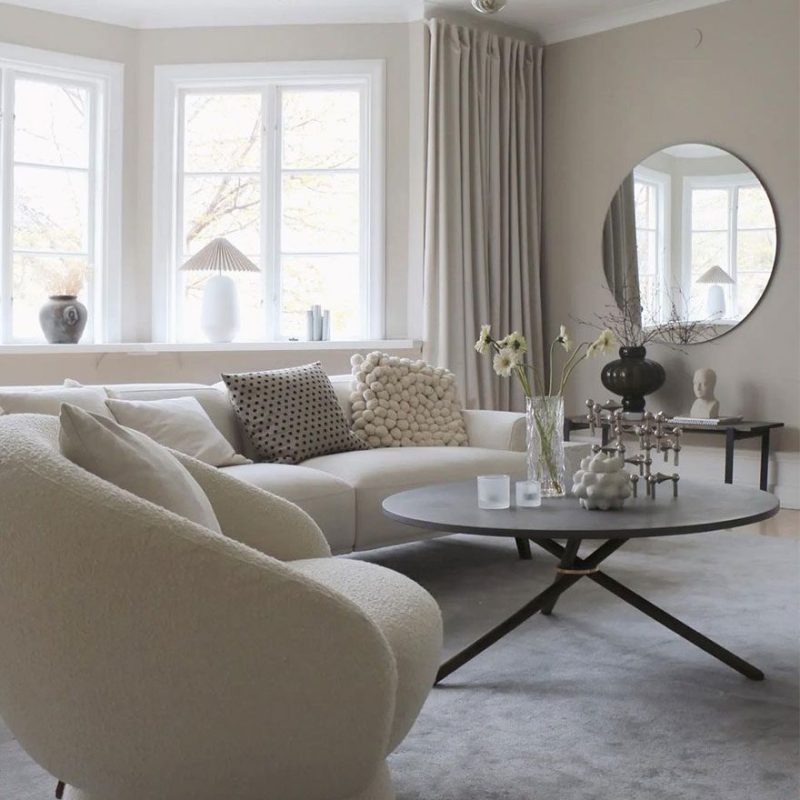 En moderne stue med neutrale farver med en plys hvid sofa, en EBERHART - Edda Sofabord Ø105 og store vinduer klædt med lange gardiner. et cirkulært spejl og en lysekrone tilføjer dekorative detaljer.