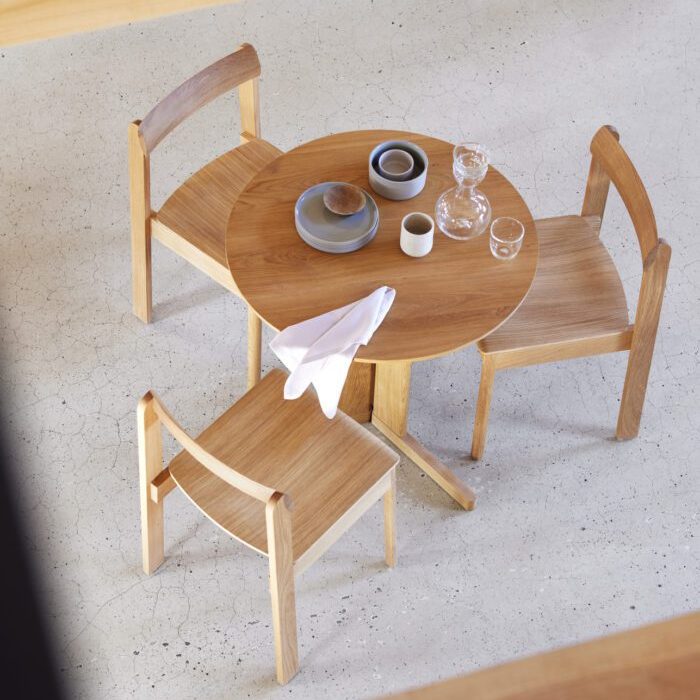 En minimalistisk spisestue med to træstole og en FORM & REFINE - Trefoil Bord Ø75. på bordet står et par fade, kopper og en spildt serviet, alt sammen under naturlig belysning.