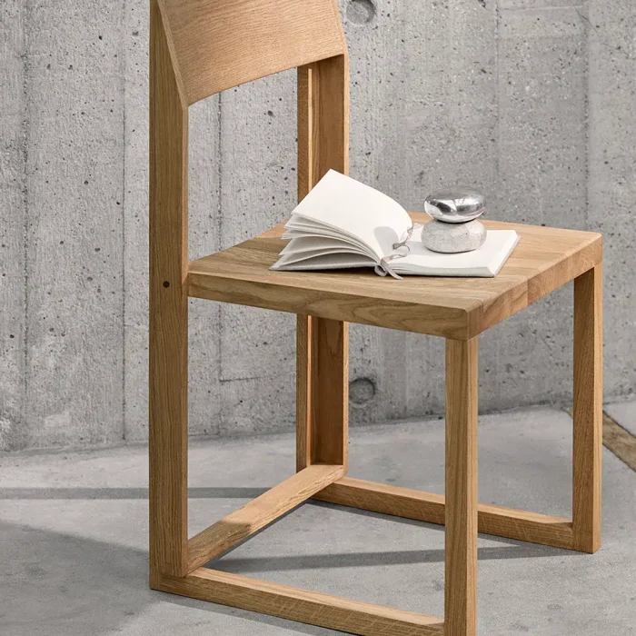En moderne træstol sat op ad en betonvæg, med et sæde med en åben bog og en lille kop på en underkop placeret på den. Stolens design inkluderer et solidt ryglæn og kantede ben, der afspejler principperne i TomorrowsDesign.