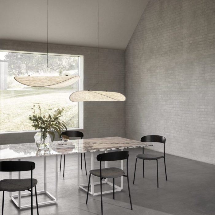 En moderne spisestue med et rektangulært bord med magasiner, omgivet af fire sorte stole, under to unikke ovale pendellamper. rummet har grå murstensvægge og et stort vindue med udsigt over grønne områder.