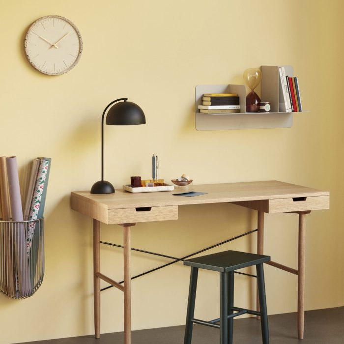 En minimalistisk hjemmekontoropstilling med et træskrivebord og matchende skammel mod en gul væg, ledsaget af en sort lampe, væghylder og et ur. Interiøret omfatter bøger, papirvarer,