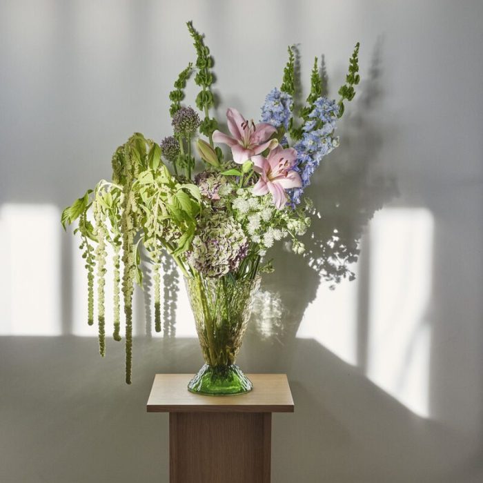 En vase med diverse blomster inklusive lyserøde liljer og grønt løv på et lille træbord, oplyst af sollys, der kaster skyggemønstre på en hvid væg bagved. Dette interiørarrangement forbedrer stemningen og afspejler sofistikeret smag i boligindretningen.